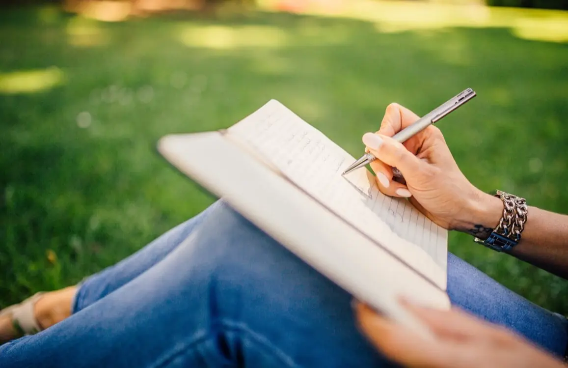 Young woman sitting in sunlit grass, jotting down her thoughts for a new book project using pen and paper.